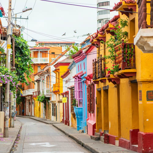 A narrow street lined with brightly colored houses in shades of yellow, blue, and pink in Cartagena, Colombia, with some greenery and balconies visible.