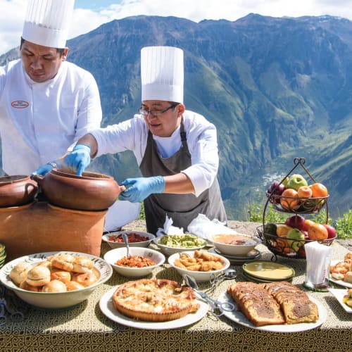Zwei Köche bereiten draußen auf einer Bergterrasse mit verschiedenen Gerichten auf dem Tisch Essen zu.