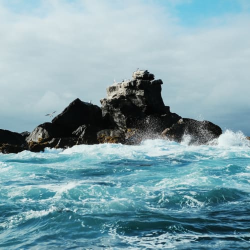 Rough ocean waves crash against dark volcanic rocks in the sea.
