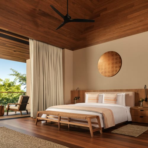 Geräumiges Hauptschlafzimmer mit Holzdecke und Holzboden, großem Bett mit weißer und beiger Bettwäsche, Bank am Fußende des Bettes, Wanddekoration, Nachttische und offener Balkon mit Blick auf Grünflächen.