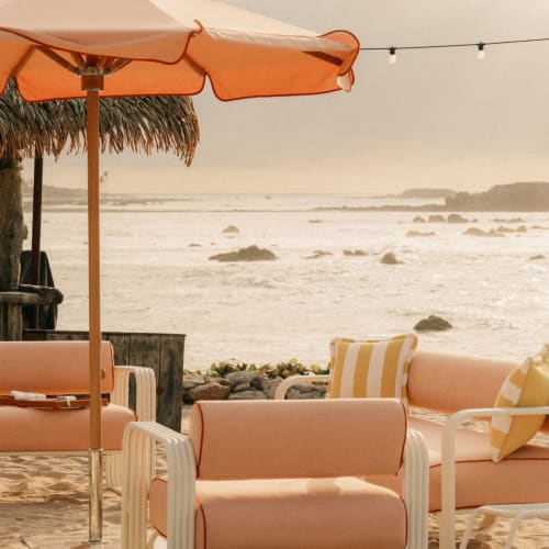 Außenbereich am Strand mit gepolsterten Sesseln, einem gestreiften Liegestuhl und einem großen orangen Sonnenschirm vor einer hölzernen Tiki-Bar, mit Blick auf felsige Küste und ruhiges Meer unter einem nebligen Himmel.