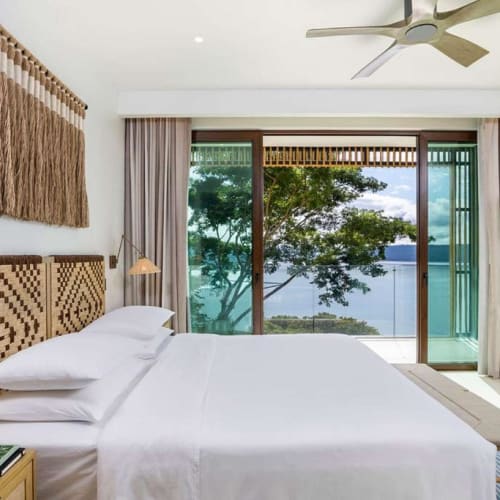A cozy hotel bedroom with a large window showcasing a view of trees and water, decorated with natural woven accents and neutral colors.