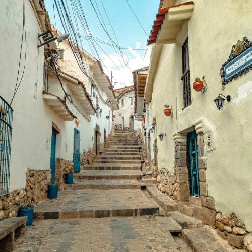A narrow stone-paved street with stairs in a quaint, historic neighborhood, lined with white buildings with blue accents, small potted plants, and decorative lamps.