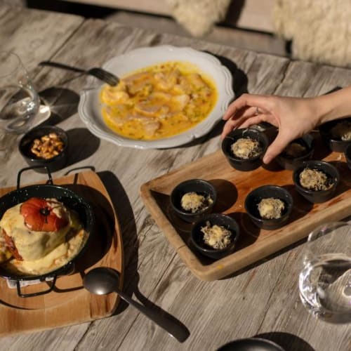 Ein Esstisch mit einem Gericht, das mit geschmolzenem Käse überzogen ist, eine Schüssel mit cremiger Suppe und mehreren kleinen Schalen mit geriebenem Käse, eine Hand greift nach einer der Schalen.
