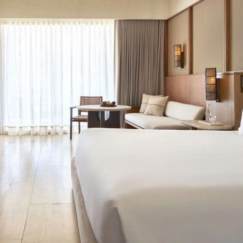 Modernes Hotelsuiten-Schlafzimmer mit großem weißem Bett, beigem Sofa, Holzmöbeln und bodentiefen, transparenten Vorhängen vor einem Fenster.