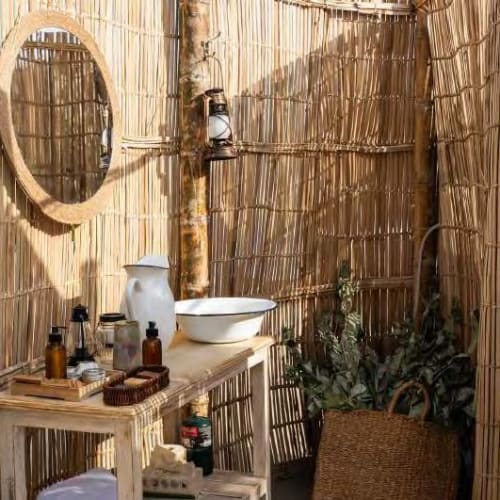 A rustic outdoor bathroom area with a bamboo fence, a mirror, and various toiletries on a small wooden table.