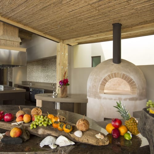 Fruits and decorative shells on a wooden counter in a modern outdoor kitchen with a pizza oven and pineapples in the background.