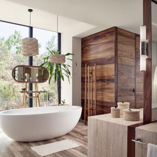 Moderne Badezimmer mit freistehender weißer Badewanne, großem Holzrahmenspiegel, hängenden Korblampen und bodentiefen Fenstern mit Blick auf das Grün draußen.