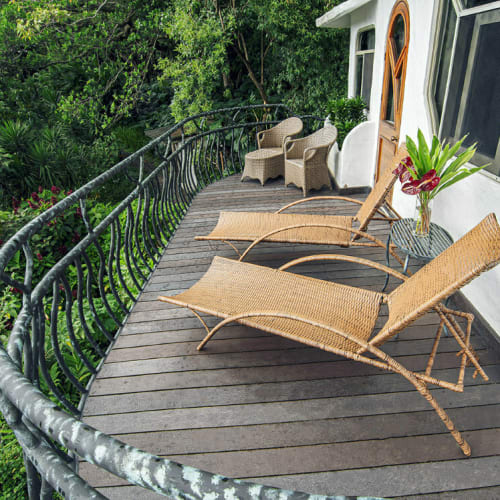 Curved wooden deck attached to a white building with two wicker lounge chairs, two wicker armchairs, a small table with a plant, overlooking dense green foliage.