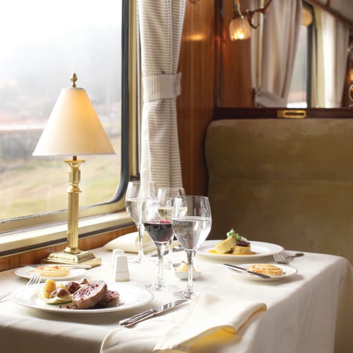 A luxurious train dining car table set with white tablecloth, elegant dishes, wine and water glasses, and a brass lamp by the window with light curtains.