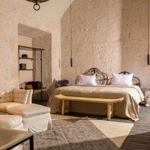 Ein gemütliches Schlafzimmer mit großem Bett, weicher Bettwäsche und einer Bank am Fußende des Bettes, vor Steinwänden mit minimalistischer Beleuchtung.