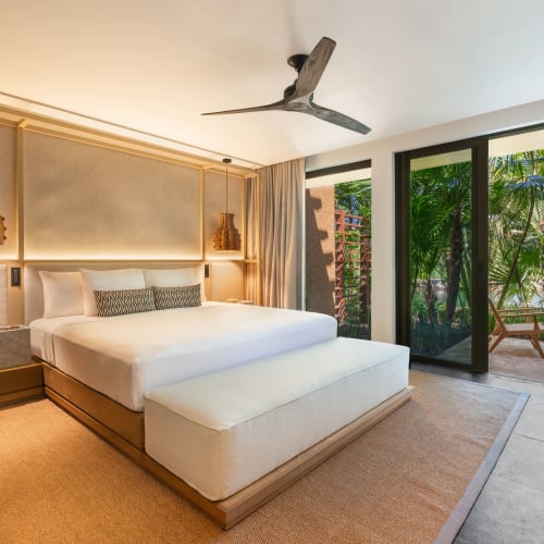 Ein modernes, luxuriöses Hauptschlafzimmer mit großem Bett mit weißer Bettwäsche, Hängeleuchten, Deckenventilator und Schiebetüren, die zu einer Terrasse mit tropischem Grün führen.