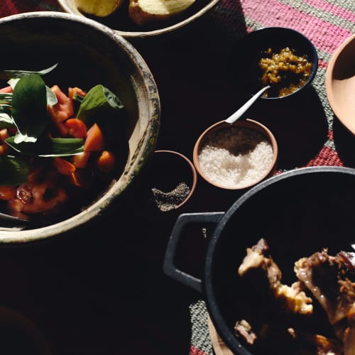 Verschiedene Gerichte, darunter eine Schüssel mit geschnittenen Kartoffeln, ein Salat mit Grünzeug und Tomaten, kleine Schalen mit Salz, Pfeffer und einem Löffel Senf oder Chutney sowie ein schwarzer Topf mit gekochtem Fleisch, alles auf einem gewebten roten und grauen Tischtuch.