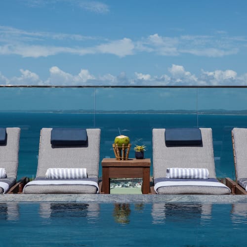 Four lounge chairs with rolled towels and headrests lined up beside a pool overlooking a blue ocean under a partly cloudy sky.