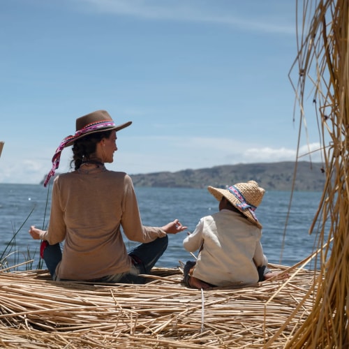 Eine Frau und ein Kind sitzen in Lotussitz auf einer Schilfplattform am See, beide tragen Hüte und blicken auf das Wasser mit einer Berglandschaft im Hintergrund.
