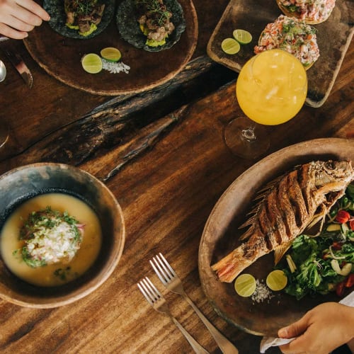 Ein Holztisch mit gegrilltem ganzen Fisch, serviert mit Salat und Limettenscheiben, einer Schüssel Suppe, zwei Gläsern mit Getränken und zwei Händen, die nach dem Essen greifen, in einem Restaurant.