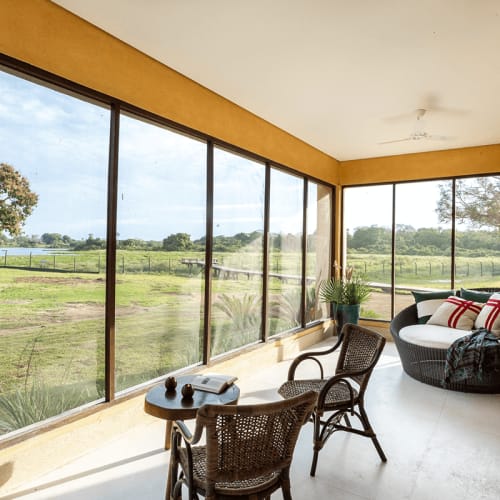 Ein heller Wintergarten mit großen Fenstern mit Blick auf eine grasbewachsene Landschaft, eingerichtet mit zwei Korbstühlen, einem kleinen Tisch mit Büchern und Dekoration sowie einer runden gepolsterten Lounge mit bunten Kissen.