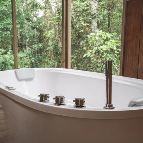 Eine moderne weiße Badewanne mit Chromarmaturen steht in der Nähe großer Glasfenster mit Blick auf einen dichten grünen Wald draußen.