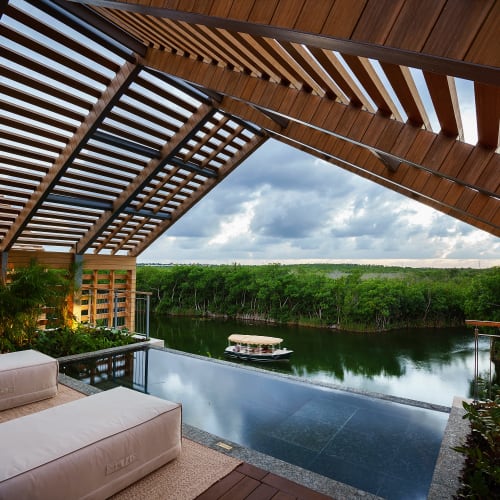 Luxuriöse Lagunen-Poolvilla mit einem Holzlamellen-Dach, weißen gepolsterten Sitzgelegenheiten und Blick auf einen ruhigen Wasserweg umgeben von üppigem Grün unter bewölktem Himmel.