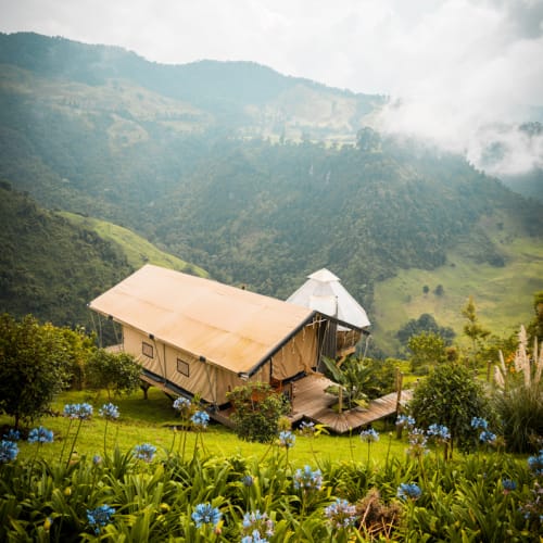 Eine Holzhütte mit beigem Stoffdach liegt eingebettet an einem grünen Hügel, umgeben von blauen Blumen und dichtem Grün, mit nebligen Bergen im Hintergrund.