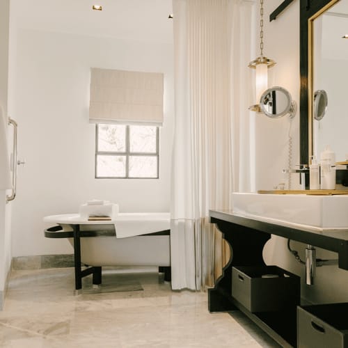 Luxusbadezimmer mit freistehender weißer Badewanne, schwarz gerahmtem Spiegel, Marmorfliesenboden und weißen Bademänteln an der Wand.