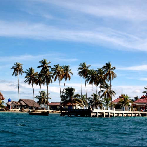 Tropische Szene mit Palmen, strohbedeckten Hütten und einer Pier am Wasser in einer Küstenregion.