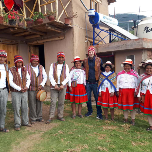 Eine Gruppe von Männern und Frauen in traditioneller Andenkleidung steht draußen vor einem rustikalen Gebäude mit Blumentöpfen und Wassertanks auf dem Dach.