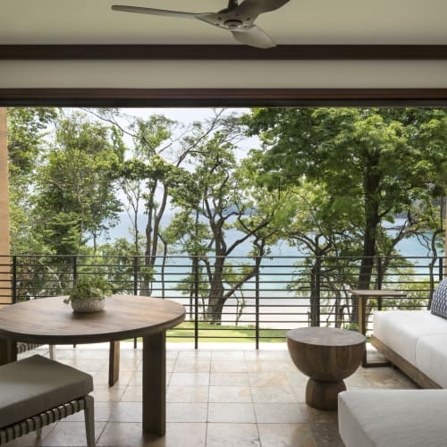 Ein geräumiger Balkon mit einem Holztisch, Stühlen und einem weißen Sofa mit Blick auf satte grüne Bäume und den Ozean.