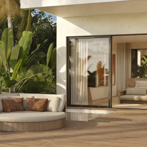 Luxuriöse Terrasse mit rundem Polstersofa und Beistelltisch, umgeben von üppigen tropischen Pflanzen, neben großen Glasschiebetüren, die zu einem modernen Wohnbereich im Inneren führen.