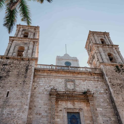 Froschperspektive auf die Fassade einer historischen Steinkirche mit zwei Türmen, eingerahmt von Palmblättern vor klarem Himmel in Valladolid, Yucatán, Mexiko.