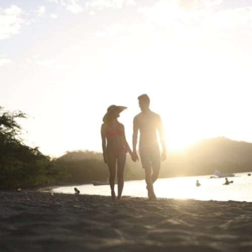 Ein Paar läuft Hand in Hand am Strand bei Sonnenuntergang, wobei die Sonne tief am Horizont eine Silhouette bildet.