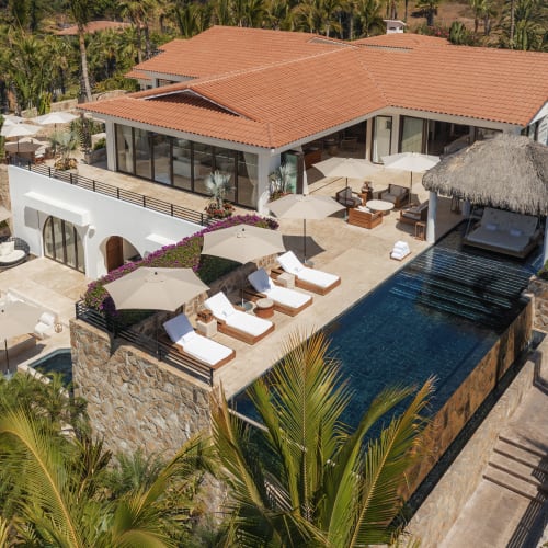 Vogelperspektive auf eine luxuriöse Villa mit einem Dach aus Terrakottaziegeln, einem Infinity-Pool und mehreren Liegestühlen, umgeben von tropischen Palmen.