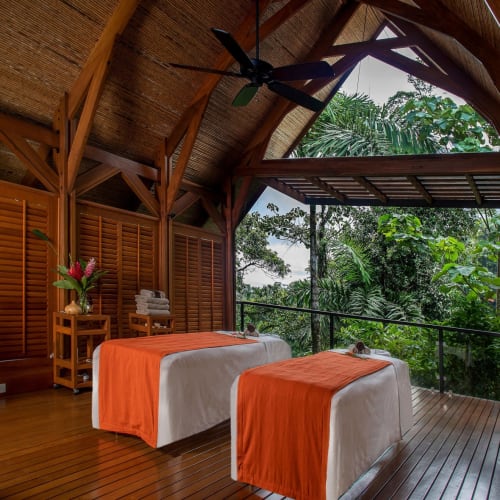 A cozy spa room with two massage tables draped in orange covers, surrounded by wooden walls and a view of lush green tropical trees.