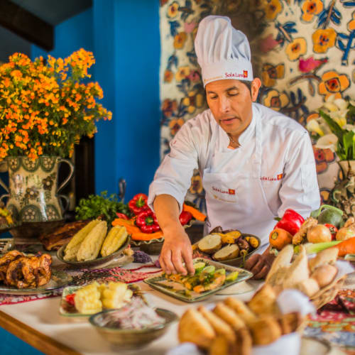 Chef in a white uniform and hat preparing a dish on a table filled with various vegetables, roasted chicken, and traditional decorations in a colorful room.