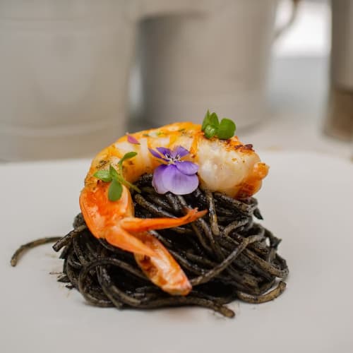 Eine Portion schwarzer Tintenfisch-Spaghetti, garniert mit einer großen gegrillten Garnele, kleinen grünen Kräutern und einer violetten essbaren Blume auf einem weißen Teller.