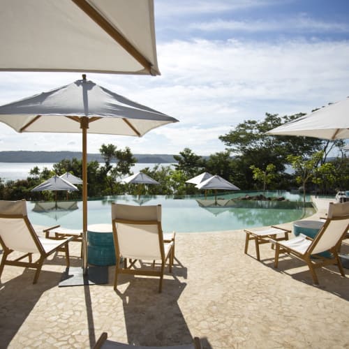 Poolseitige Liegestühle mit Sonnenschirmen, die einen Swimmingpool und einen malerischen Seeblick im Hintergrund überblicken.