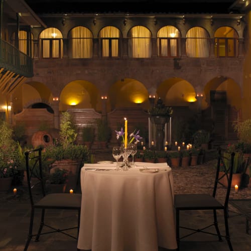 A romantic dinner setup for two with a round table draped in white cloth, candles, and purple flowers, set in a historic courtyard at night with warm lighting and lush plants.