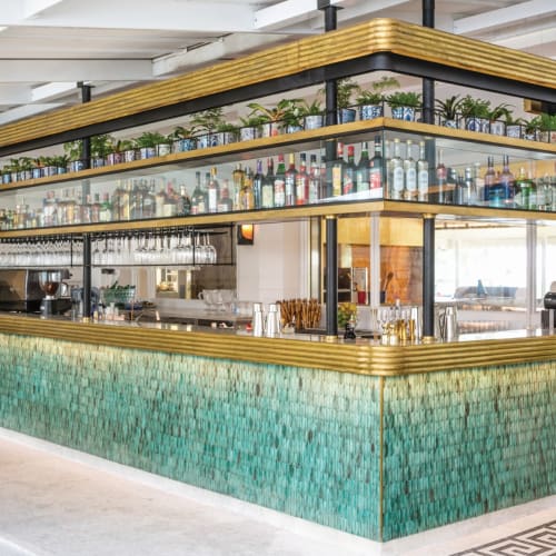 A stylish bar with a golden-edged counter and multiple shelves of bottles and plants inside a modern restaurant.