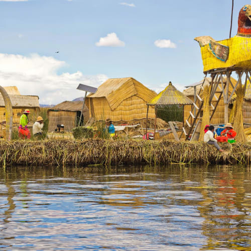 A lakeside village with traditional thatched huts and large yellow bird-shaped structures made of reeds, with people engaged in daily activities near the water under a partly cloudy sky.