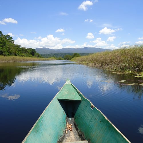 A green boat is floating on a calm river with lush greenery on both sides and a mountain range in the distance under a partly cloudy sky.