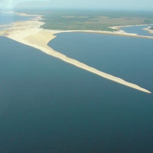 Luftaufnahme eines Flusses, der auf eine Wasserfläche trifft, mit Sandbänken und grünem Land in der Ferne.