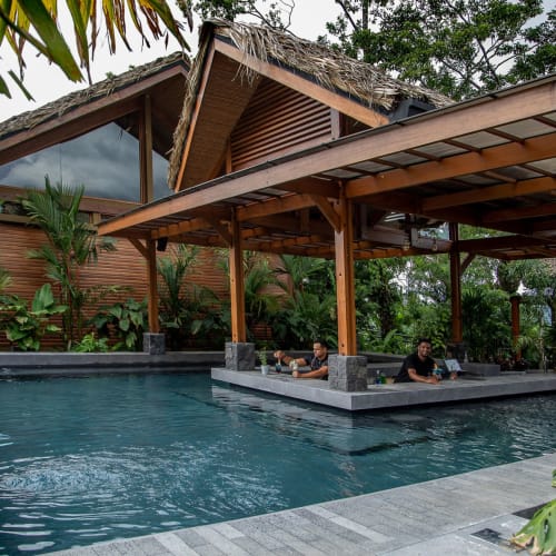 Ein Swimmingpool mit einem Holzgebäude und einer Bar in einer tropischen Umgebung, umgeben von üppigem Grün.