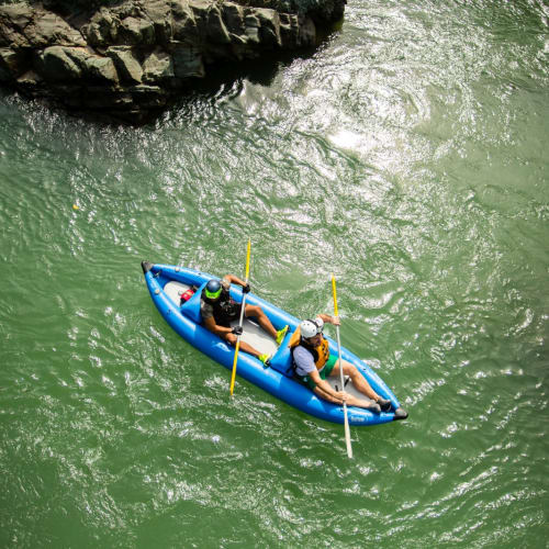 Zwei Personen in einem blauen offenen Kajak paddeln auf einem Fluss, umgeben von Felsen und Grün.