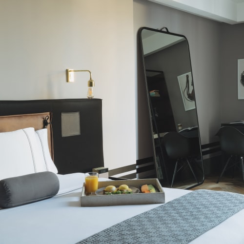 Modernes Hotelzimmer mit großem Bett, grauen Zylinderkissen, einem Tablett mit Frühstücksessen, einem Standspiegel und einem schwarzen Schreibtisch mit Stuhl in der Ecke.