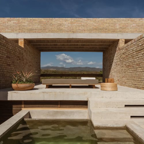 Ein moderner Außenbereich mit rechteckigem Pool im Vordergrund, Betontreppen, einem niedrigen Bett mit Kissen und Topfpflanzen, umgeben von Ziegelmauern unter blauem Himmel.