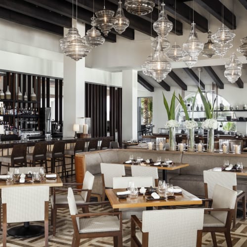 Heller und moderner Restaurantinnenraum mit Holzbalken, hängenden Glaspendelleuchten, Barbereich und Tischen mit Geschirr und weißen Stühlen.