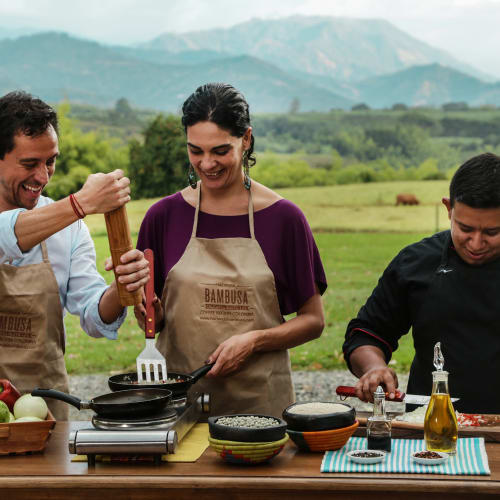 Three people participating in an outdoor cooking class in a lush green landscape with mountains in the background, smiling and cooking together.
