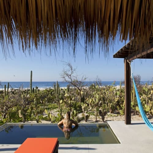 Eine Frau entspannt in einem privaten Pool mit Blick auf eine Wüstenlandschaft mit Kakteen und das Meer im Hintergrund, unter einem Strohdach und einem hölzernen Pergola mit einer blauen Hängematte.