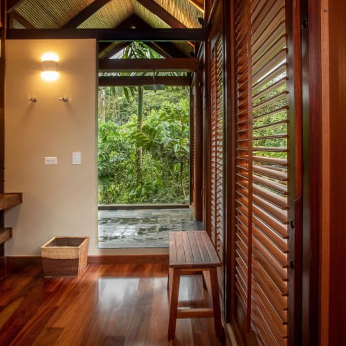 A cozy bathroom with wooden shutters and a view of lush greenery outside, featuring a sink with a mirror and towels.