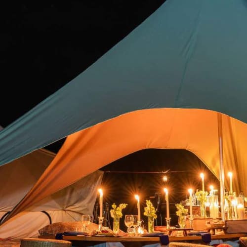 Eine gemütliche Essensstelle in einem großen Zelt am Strand bei Nacht, beleuchtet von Kerzen und umgeben von Kissen.